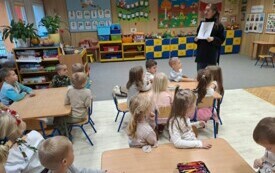 Dzieci przy stolikach obserwują prowadzącą wyjaśniającą pracę plastyczną