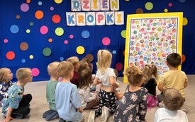 DZIECI SIEDZĄ NA DYWANIE PRZED TABLICĄ I PRZYGLĄDAJA SIĘ KOLOROWEJ PLANSZY